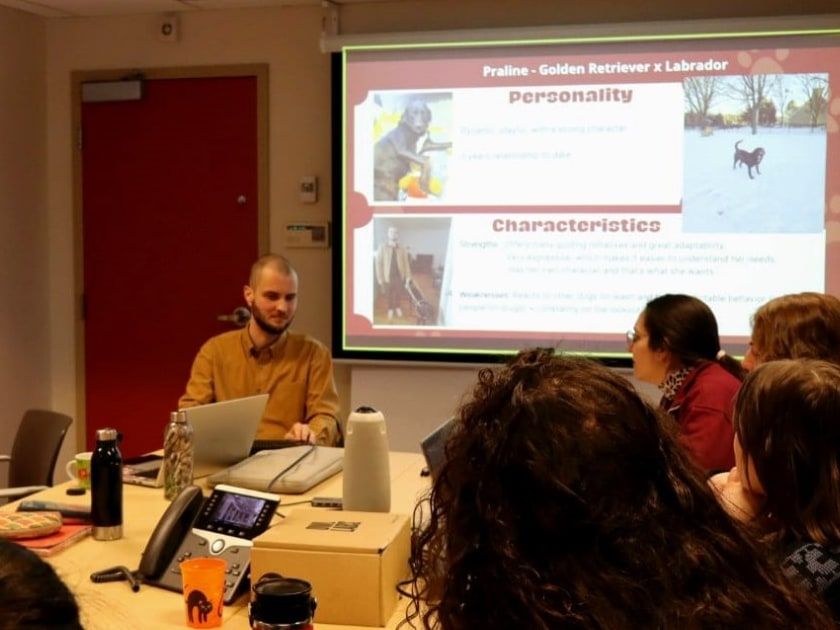 Marc de Montlivault donne une conférence, assis au bout d’une table de réunion devant son ordinateur portable. Derrière lui, une diapositive présente un chien-guide. Au premier plan, des participantes l’écoutent, vues de dos.
